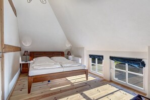 Ein Schlafzimmer mit einem hölzernen Bett, einem Nachttisch, einer Lampe und Blick ins Freie durch ein Fenster.