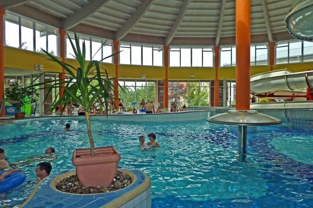 An indoor swimming pool with multiple lanes, a central water slide, and a seating area with a view of the outdoors.