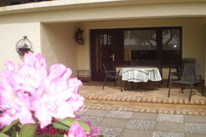 Un espace extérieur aménagé en terrasse, avec une table et des chaises, un pot de fleurs et une porte donnant sur l’intérieur.