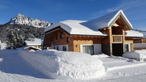 Ein Chalet-Stil Gebäude mit geneigtem Dach, umgeben von verschneiterten Flächen und Bergen im Hintergrund.