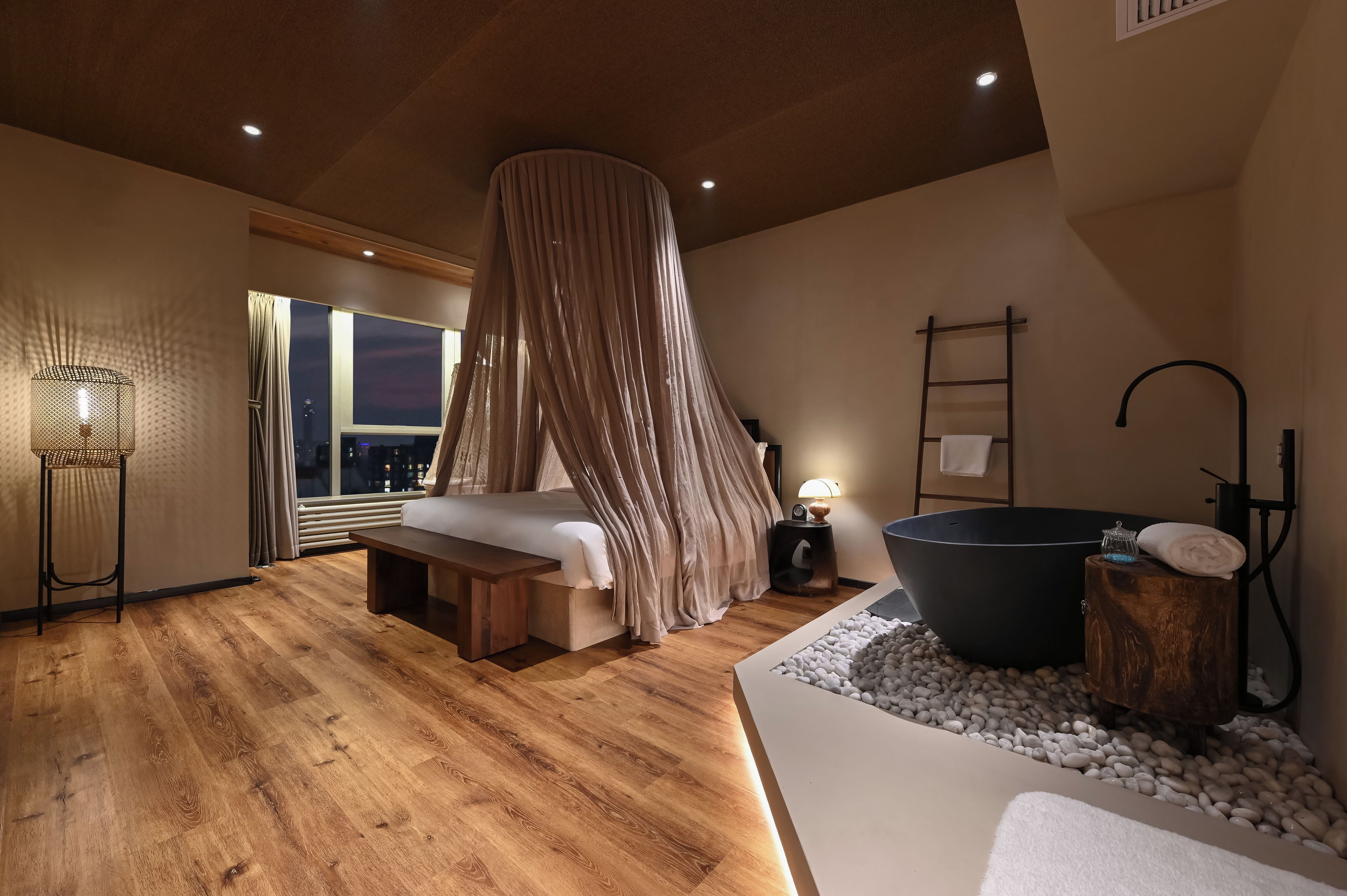 A modern bedroom with a large bed, a black bathtub, and a view of the city at night.