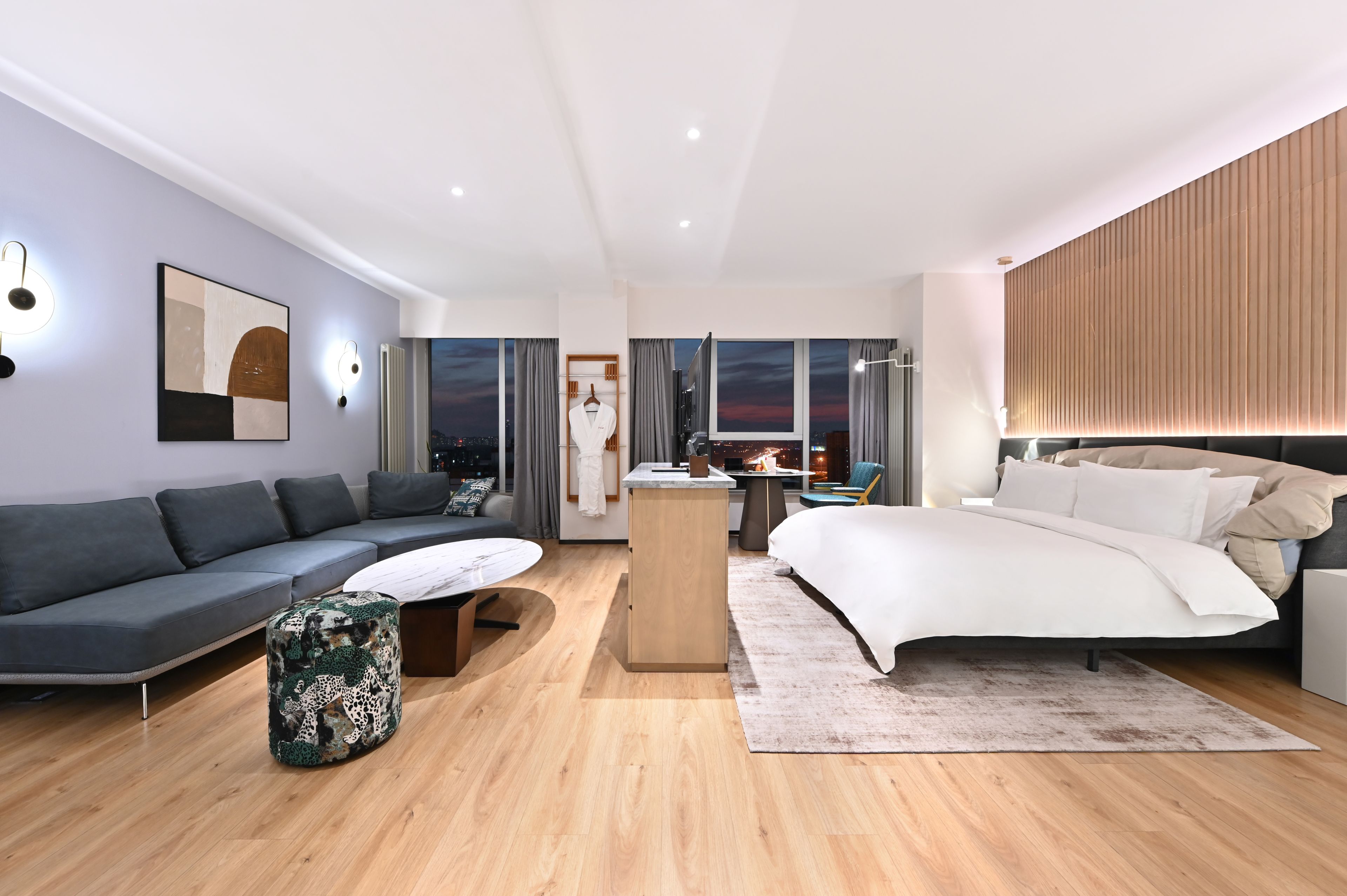 Una habitación de hotel moderna con sofá, cama, una mesita y vistas a la ciudad al atardecer.