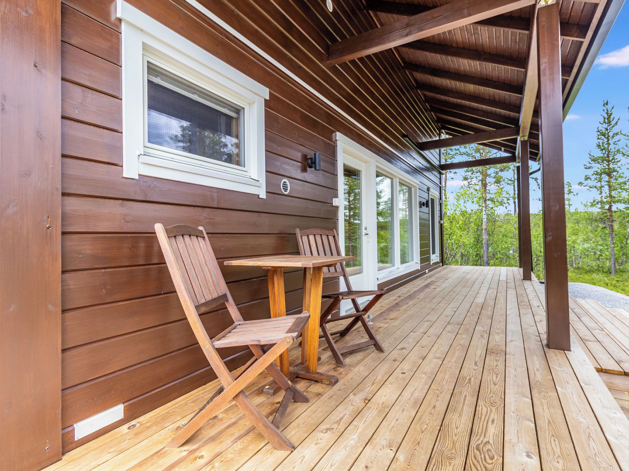 Eine Holzterrasse mit zwei Klappstühlen und einem Tisch, ein überdachter Veranda-Bereich und Blick auf Bäume.