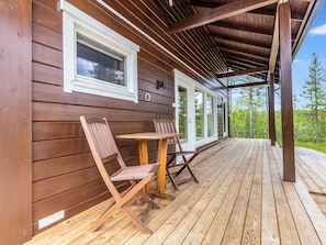 Eine Holzterrasse mit zwei Klappstühlen und einem Tisch, ein überdachter Veranda-Bereich und Blick auf Bäume.