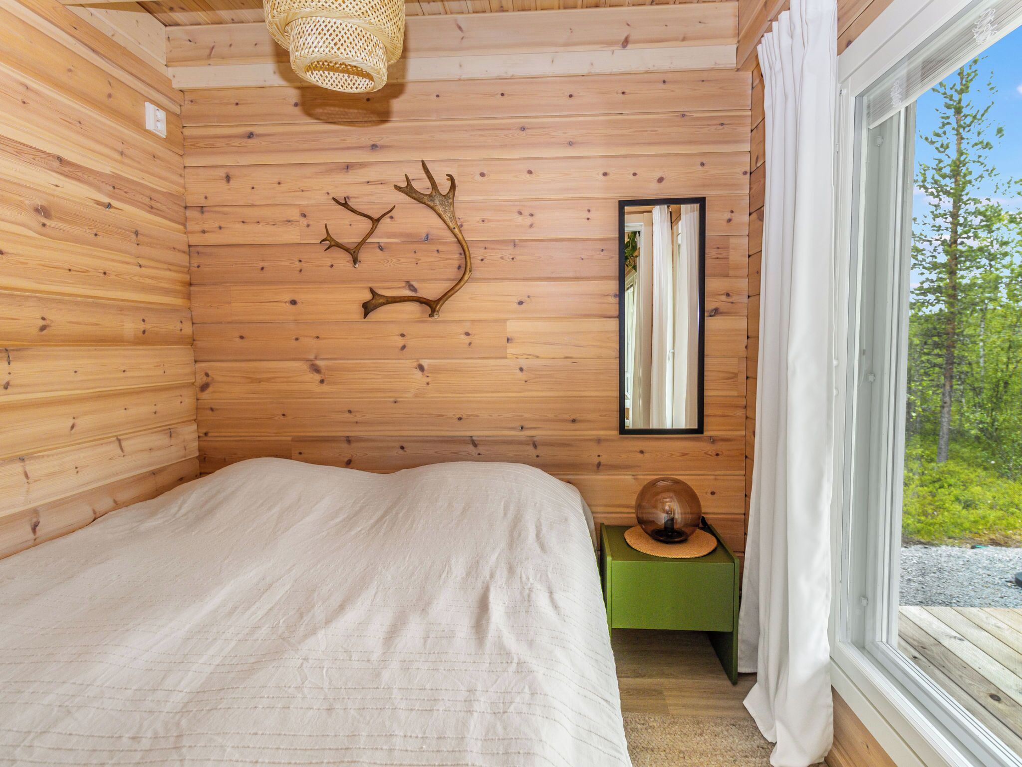 Ein Zimmer in einer Holzhütte mit einem Bett, einem Spiegel, einem Wandbrett mit Hirschgeweih und einem Fenster mit Blick auf Grünflächen.