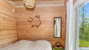 A wooden cabin room with a bed, a mirror, a wall-mounted deer antler, and a window with a view of greenery. - Villa goskanjavri by Interhome (Inari)
