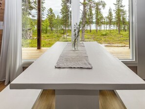 Ein modernes Interieur mit einem großen weißen Tisch, einer Pflanze in einer Vase und Blick auf einen Wald durch große Fenster.