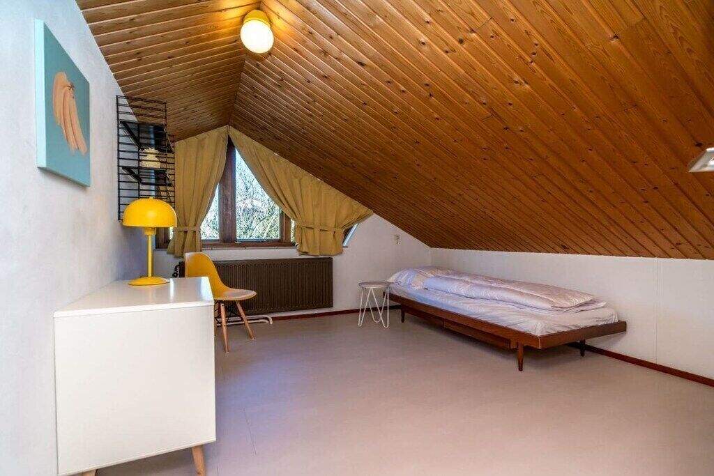 Une petite chambre à coucher de style mansardé, avec un plafond en pente, un lit simple, un bureau avec une chaise et une fenêtre avec des rideaux.