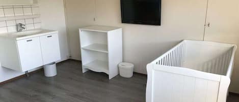 A room with a white crib, a white cabinet, a white shelf, and a television on the wall.