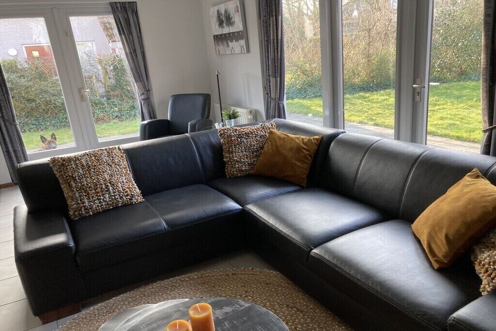 Een zwarte leren bank met gekleurde kussens, een transparante glazen salontafel en uitzicht op een tuin door grote ramen.