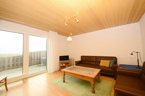 A living room with a glass-top coffee table, a brown leather sofa, a television, and a view through large sliding glass doors. - "Kachelot" in the Nixe House (Juist)
