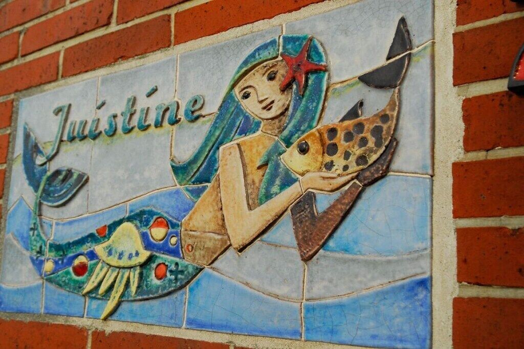 A mosaic with a mermaid and fish, and the word 'Justine' on a brick wall.