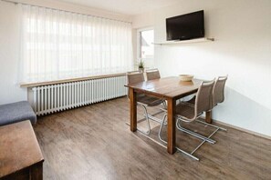 A room with a wooden dining table, chairs, a television mounted on the wall, and a window with sheer curtains. - Holiday apartment in Juist with swimming pool (Juist)