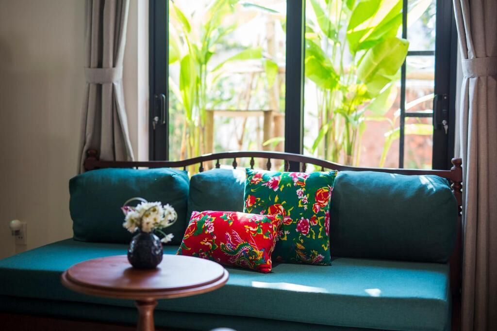 A green sofa with floral cushions, a small table with a vase, and a view of greenery outside.