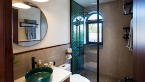 A modern bathroom with a round mirror, a green sink, a glass-enclosed shower, and a window providing natural light. - La Maison Gao (Hoi An Dong ward)