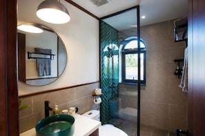 A modern bathroom with a round mirror, a green sink, a glass-enclosed shower, and a window providing natural light. - La Maison Gao (Hoi An Dong ward)