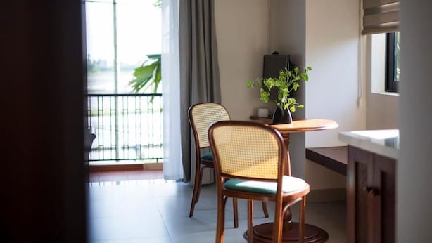 Habitación con una mesa de comedor, dos sillas de mimbre, una planta en maceta y vista a un balcón con barandilla.