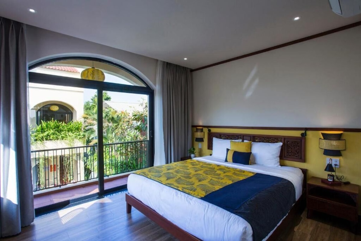 A hotel room with a large bed, a nightstand, a lamp, and a view of a balcony with plants.