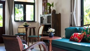 Una habitación con un sofá verde, una silla de madera, una mesita redonda con un florero y una ventana con cortinas.