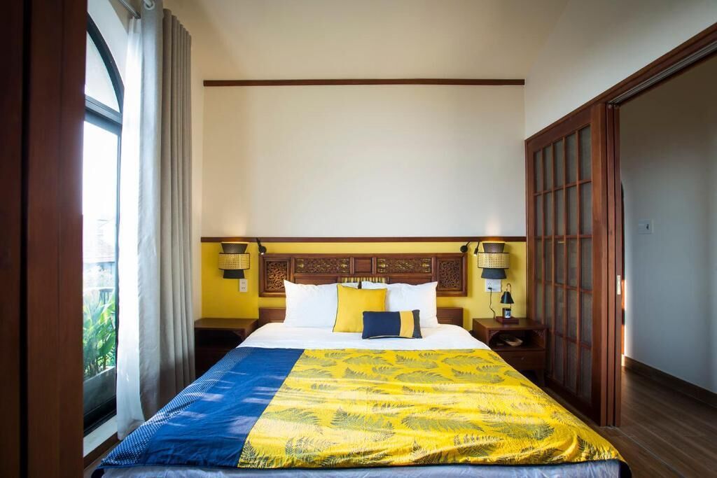 A hotel room with a large bed, a wooden headboard, two bedside tables with lamps, and a view of greenery through the window.