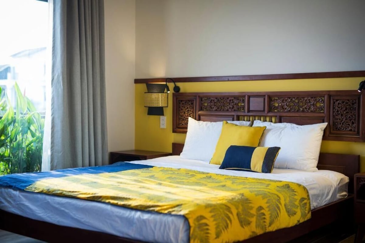 A hotel room with a bed, a nightstand, a lamp, and a view of greenery outside.