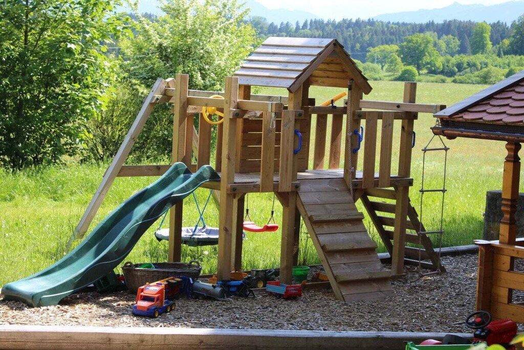 A wooden play structure with a slide and climbing area, surrounded by a grassy field and trees.
