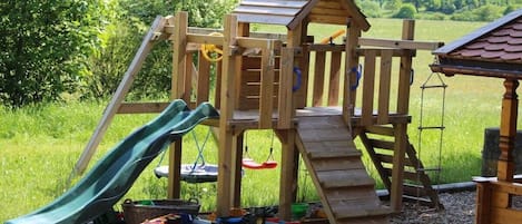 A wooden play structure with a slide and climbing area, surrounded by a grassy field and trees.
