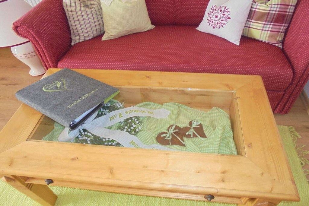 A wooden chest with a green cloth inside, placed on a wooden table in a room with a red sofa and patterned cushions.