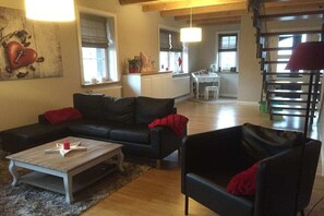 A living room with a black sofa, a coffee table, and a red throw pillow. There is a dining area with a table and chairs in the background. - Holiday home with 2 bedrooms (Letschin OT Gross Neuendorf)