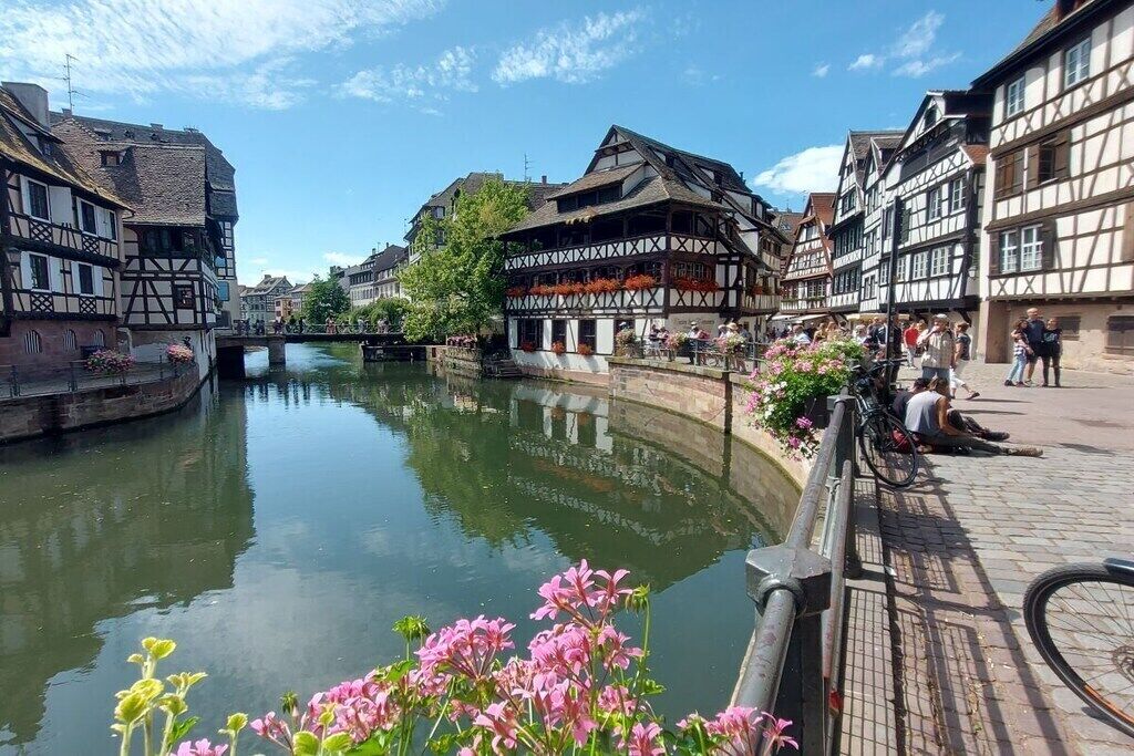 A riverside walkway with traditional half-timbered buildings, a bridge, and people walking.