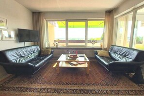 Sala de estar con dos sofás de cuero negro, una mesa de centro de vidrio, un televisor de pantalla plana montado en la pared y un ventanal con vistas a zonas verdes.