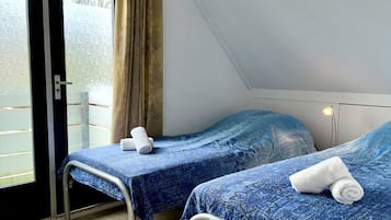 Habitación con dos camas, cada una con colcha azul estampada y toallas blancas. Se ve una ventana con cortina, y el techo es inclinado.