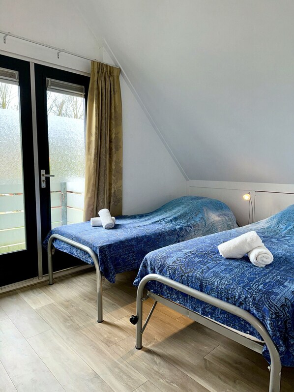 A room with two beds, each with a blue patterned bedspread and white towels. There is a window with a curtain, and the room has a sloped ceiling.