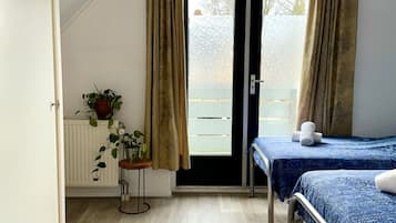 Una habitación con techo inclinado, un par de puertas dobles que dan al exterior, dos camas con ropa de cama azul, un radiador, una mesita con una planta y una planta en maceta en el suelo.
