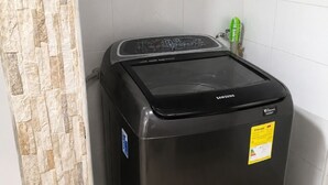 A black Samsung washing machine in a corner of a room with tiled walls and a stone-textured partition. - 3-Bedroom Family Apartment with Pool in Yopal (Villavicencio)