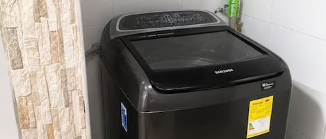 A black Samsung washing machine in a corner of a room with tiled walls and a stone-textured partition.
