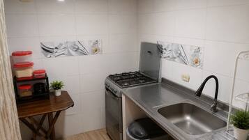 A compact kitchen with white tiles, a stainless steel sink, a stove, and a trash bin.