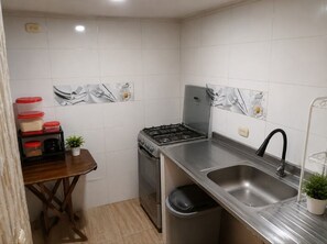 A compact kitchen with white tiles, a stainless steel sink, a stove, and a trash bin.