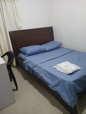 A single bed with blue bedding, a wooden headboard, a white towel on the bed, and a black chair to the left.
