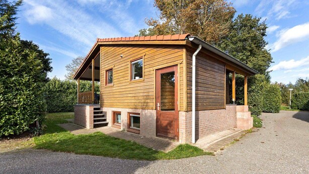 A wooden cabin with a brick foundation, a covered porch, and a gravel driveway. - Wighenerhorst Bungalow Park - 94 (Wijchen)