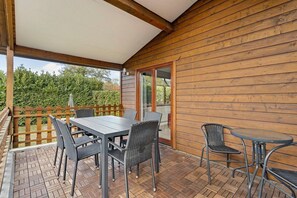 Een overdekt terras buiten met een tafel en stoelen, houten wanden en uitzicht op groen.
