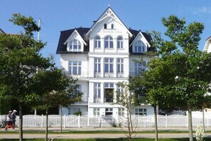 Ein großes, weißes, mehrstöckiges Gebäude mit einer symmetrischen Fassade, einem zentralen Eingang und einem Balkon im zweiten Stock.