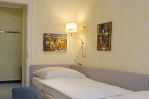 Ein Einzelbett mit weißer Bettwäsche in einem Zimmer mit cremefarbenen Wänden. An der Wand hängt eine Wandleuchte und zwei gerahmte Bilder.