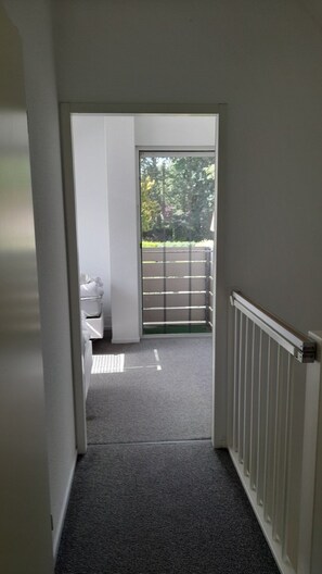 A hallway leading to a balcony with a view of trees and a fence.