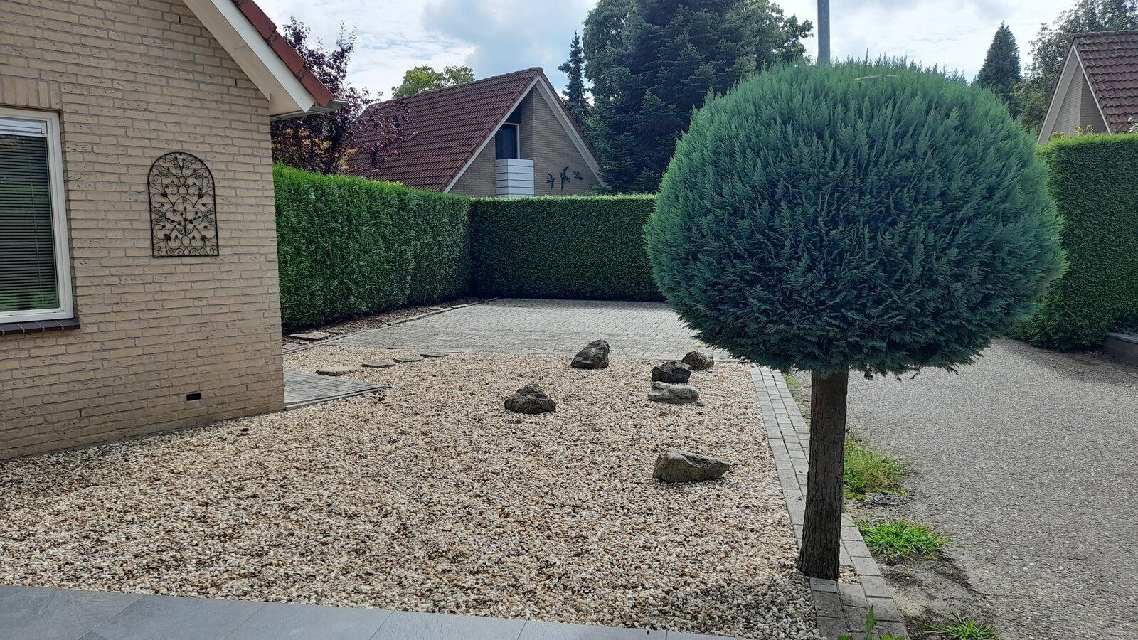 Een woonwijk met een grindpad, een strakke haag en een kleine boom.