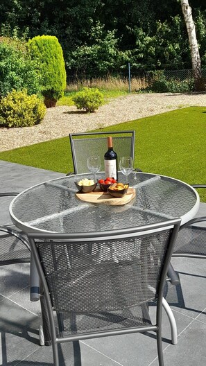 Een ronde tafel met glazen blad, metalen stoelen, een fles wijn en schalen met fruit en lekkernijen op een terras.