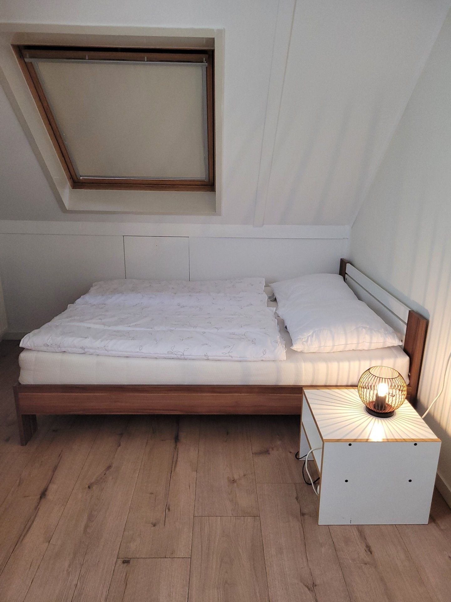 Een kleine kamer met een enkel bed, schuine balken, een raam en een nachtkastje met een lamp.