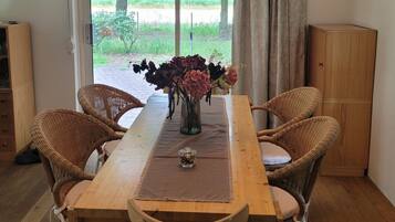 Un comedor con mesa de madera, sillas de mimbre, un florero con flores y vistas a través de un ventanal.
