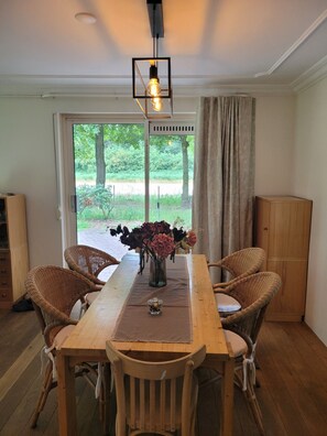 Et spiseområde med et træbord, fletstole, en vase med blomster og udsigt gennem en glasdør.