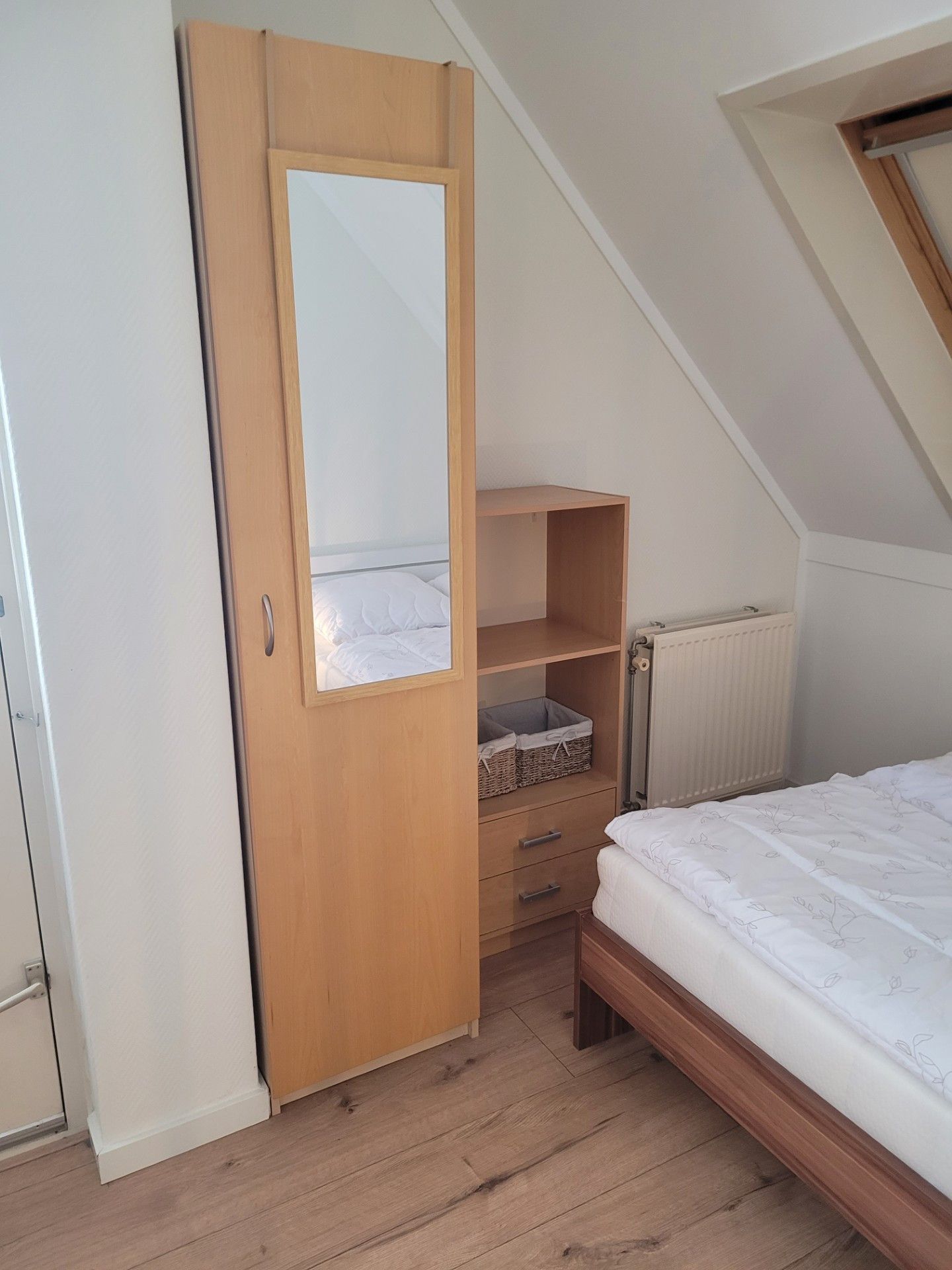 Une chambre avec une armoire en bois, un lit recouvert d’une couette blanche et un radiateur.
